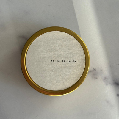 Round gold candle with 'fa la la la la...' text on a marble surface