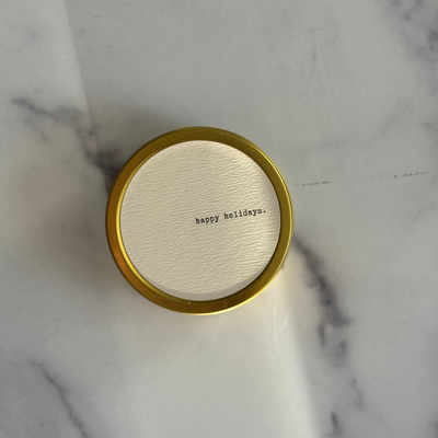 Round gold container with 'happy holidays.' text on a marble surface
