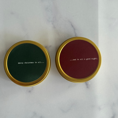 two travel tin candles with christmas inspired greetings on a marble surface.